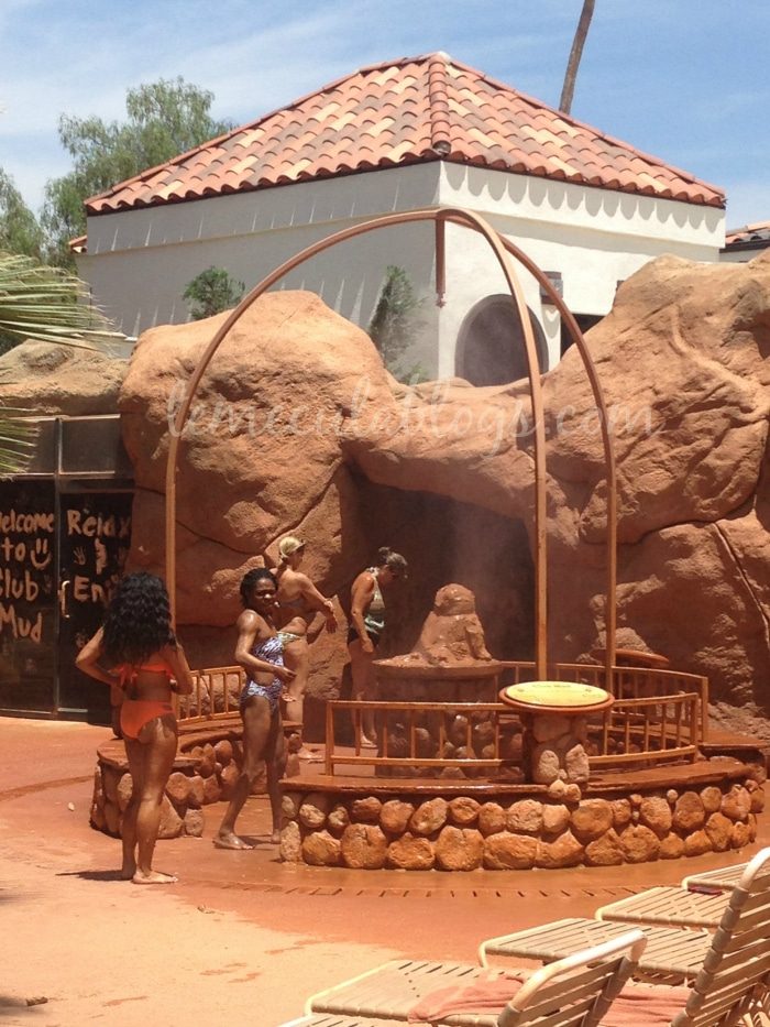 People in swimsuits stand around a mud bath area with rock formations and a wooden structure. In the background, there is a building with a terracotta-tiled roof. A sign reading "Welcome to Club Mud" is visible. A few lounge chairs are in the foreground, showcasing one of the best spas in Southern California.
