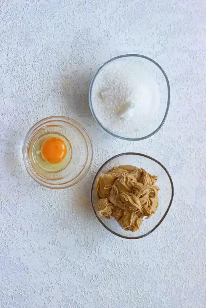A top-down view of three bowls on a textured white surface: one with a raw egg and yolk, another with granulated sugar, and the third containing creamy peanut butter—solid for whipping up 3 ingredient cookies.