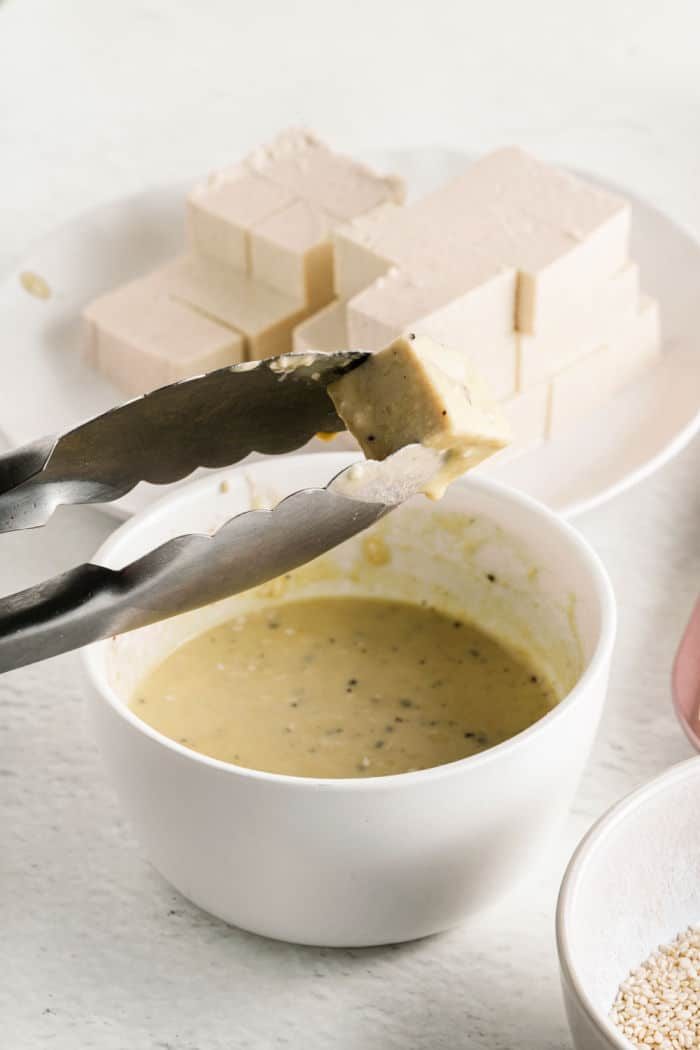 Tongs holding a tofu cube over a bowl of yellow sauce resemble the careful preparation of tofu nuggets, with more tofu blocks waiting on a plate in the background.