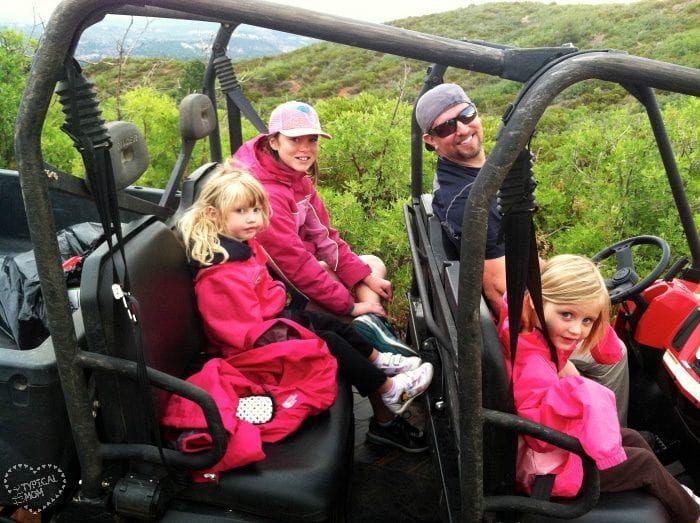 A man and three children sit in an off-road vehicle on a dirt path surrounded by green bushes and hills, embodying the spirit of travel for free. They are all wearing casual outdoor clothing, ready to embrace their spontaneous adventure.