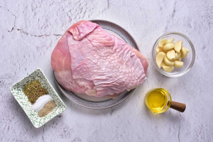 A raw turkey breast on a plate is placed on a kitchen countertop. Surrounding it are a small dish of spices, including salt and pepper, a bowl with peeled garlic cloves, and a small container of olive oil—solid ingredients for preparing Turkey Confit.