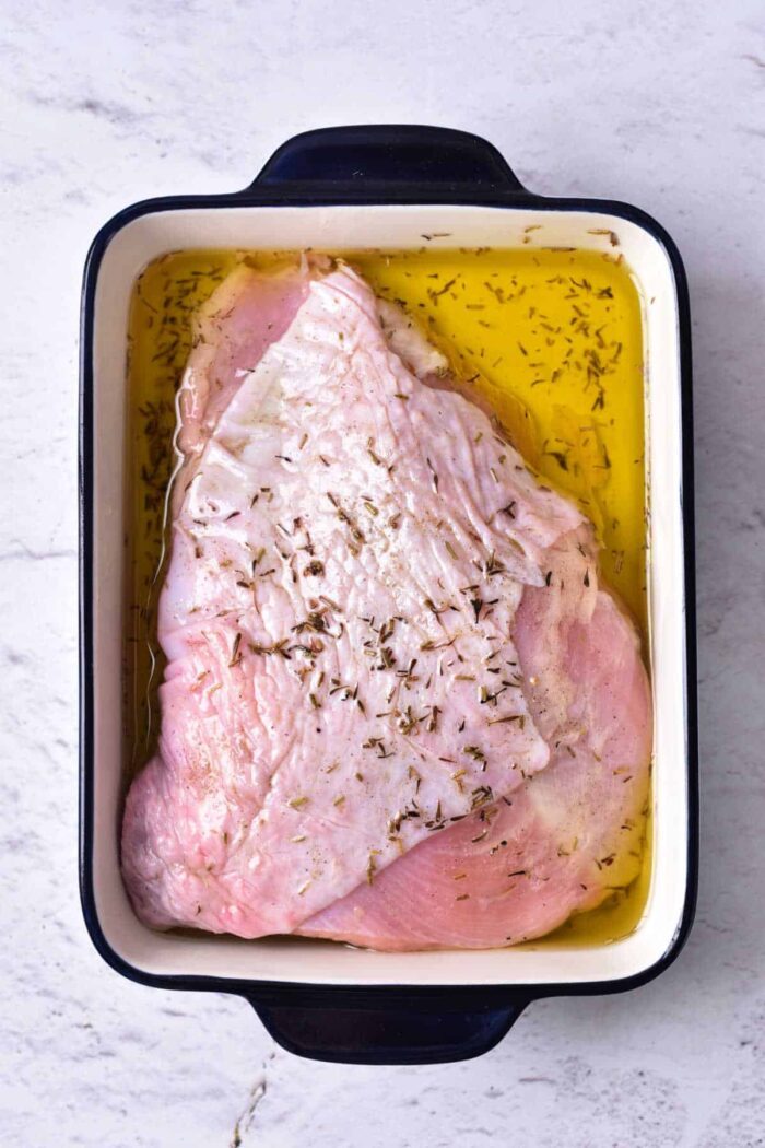 A raw turkey breast marinating in oil and seasonings, destined for a good turkey confit, sits in a rectangular white and black roasting pan on a light-colored surface.