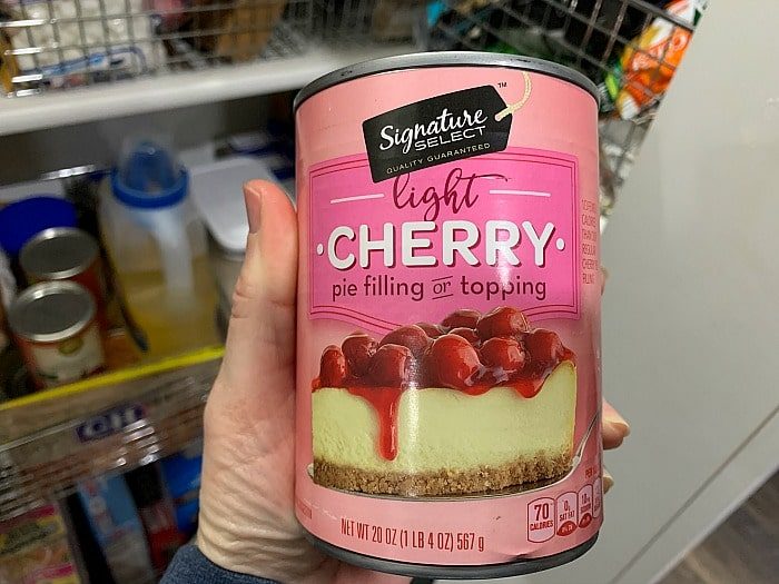 A hand holds a can of Signature Select light cherry pie filling or topping, solid for adding a fruity twist to your cherry brownies. The label showcases cherries tantalizingly perched on a cheesecake, while shelves stocked with various grocery items create a vibrant backdrop.