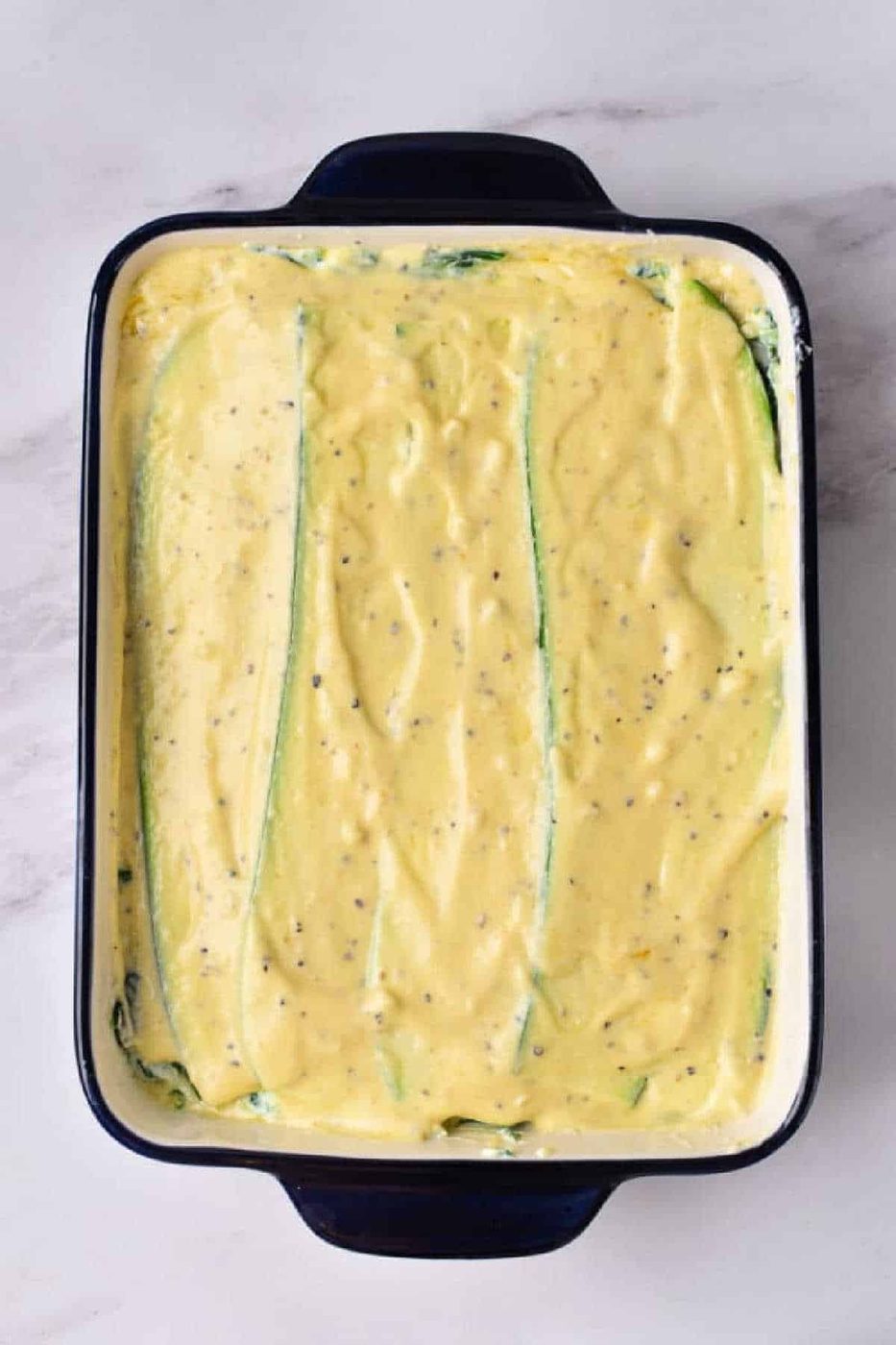 Rectangular baking dish with creamy yellow sauce spread over zucchini slices, ready for baking—this Zucchini Lasagna with White Sauce promises a deliciously comforting twist on a classic favorite.