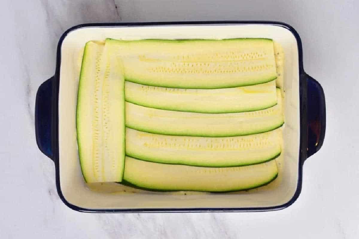 Rectangular dish with thin, raw zucchini slices neatly layered inside, ready to become a good vegetable lasagna with white sauce.