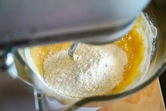 A close-up view of a stand mixer with a dough hook attachment, mixing flour and yellow batter in a metal bowl. Some dry flour is still visible on top, hinting at the transformation into good blueberry muffin tops.