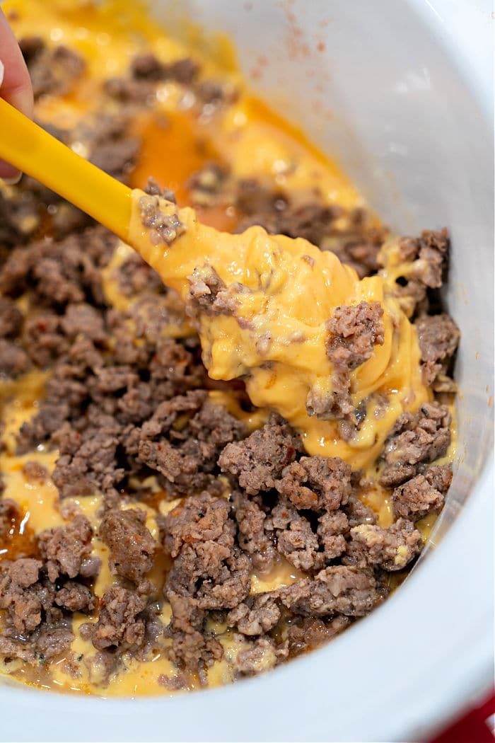 Melted Velveeta cheese and browned ground spicy sausage are being stirred in a slow cooker with a yellow spatula, creating a good Sausage Dip.