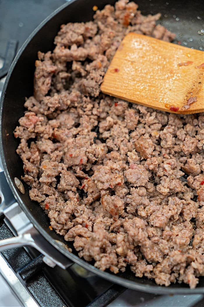 Ground sausage sizzles in a black skillet on the stovetop, stirred gently with a wooden spatula, creating a solid base for a hearty Velveeta and Rotel dip.