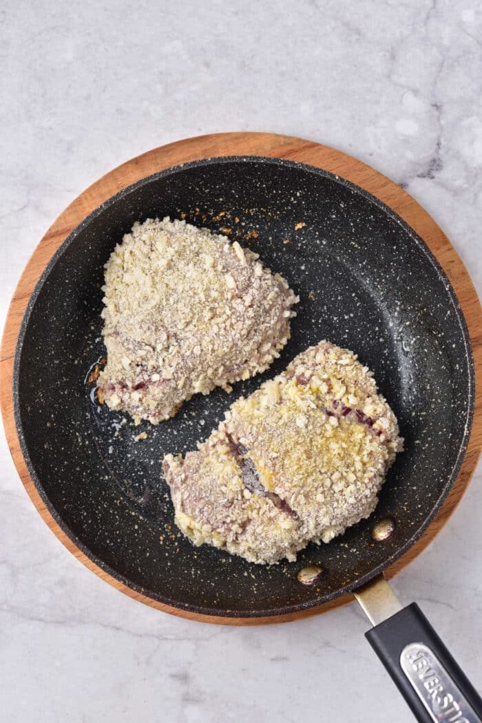 Two breaded venison steaks are sizzling in a non-stick frying pan on a marble surface in process for crafting a solid venison cube steak recipe.