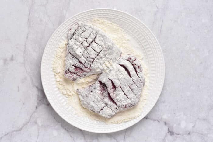 Two raw, flour-dusted deer steak rest on a white plate, placed on a marble surface