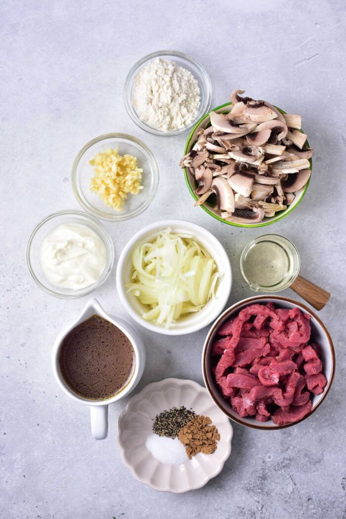 Ingredients for Venison Stroganoff in bowls: sliced venison, mushrooms, onions, garlic, flour, yogurt, oil, broth, and spices.