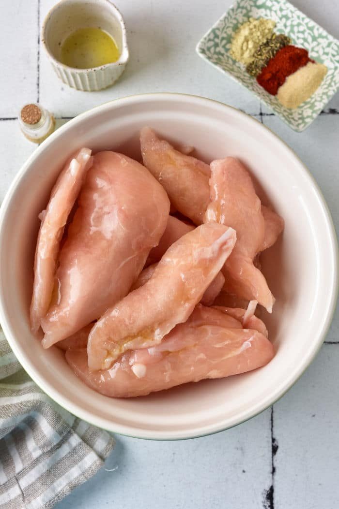Raw chicken tenderloins, sit in a white bowl beside oil and an array of spices on a tiled countertop.