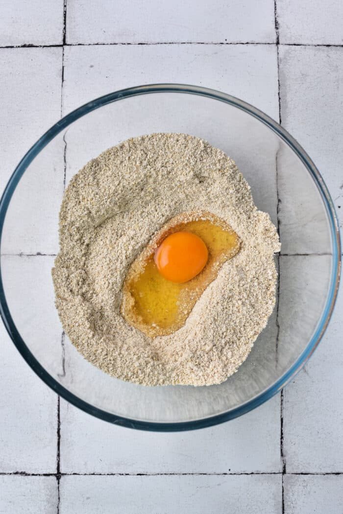 A glass bowl with matzo and an egg in the center sits on a tiled surface, reminiscent of the beginnings of a matzo ball preparation.