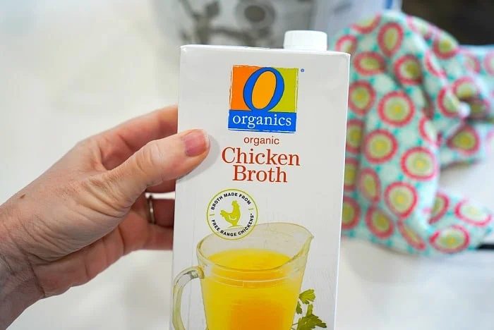 A hand holds a carton labeled O Organics Organic Chicken Broth, solid for your Instant Pot recipes. The carton showcases an image of yellow broth in a clear pitcher with a parsley garnish. A patterned cloth provides a charming backdrop.
