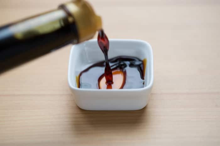 A small stream of coconut aminos are like soy sauce, an ancient and savory condiment, cascades seamlessly into a pristine white dish on a wooden surface.