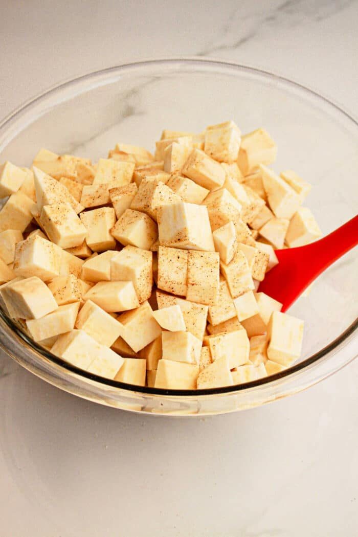 Glass bowl of cubed raw sweet potato with seasoning, stirred by a red spatula on a white countertop—solid for adding to your favorite White Sweet Potato Recipe.