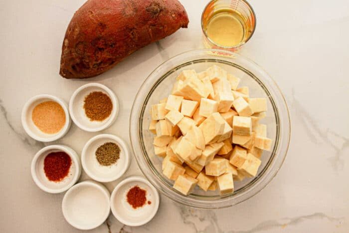 Cubed white sweet potatoes in a bowl, a whole white sweet potato, oil, and spices in small bowls on a white surface—solid for your next good White Sweet Potato Recipe.
