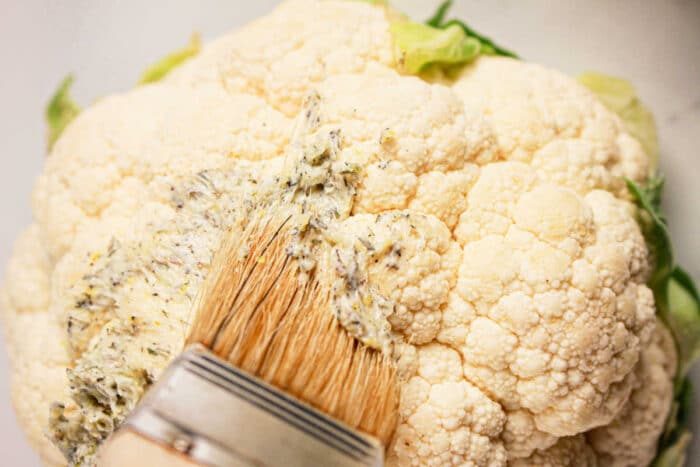 Close-up of a whole cauliflower being brushed with seasoned oil or butter using a flat brush, solid for a crock pot transformation.
