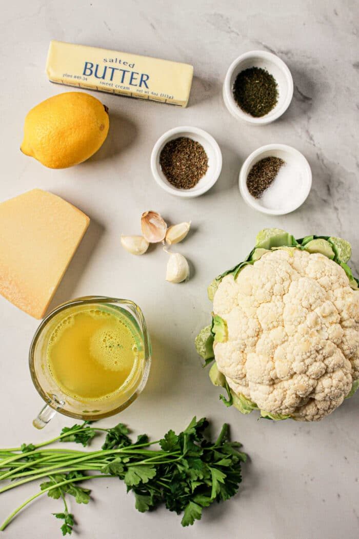 Ingredients on a marble surface: butter, lemon, cheese, garlic, whole cauliflower, herbs, broth in a jug, and small spice bowls. solid for creating a delightful cauliflower recipe that can easily be adapted for Crock Pot cooking.