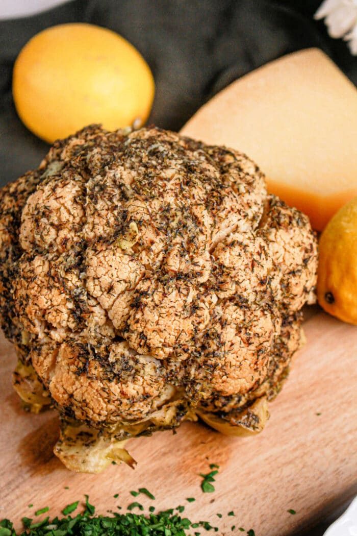 A whole roasted cauliflower, seasoned with herbs, sits on a wooden board surrounded by lemons and cheese. This delightful recipe showcases the versatility of this humble vegetable, solid for savoring as is or prepared in a crock pot for tender perfection.