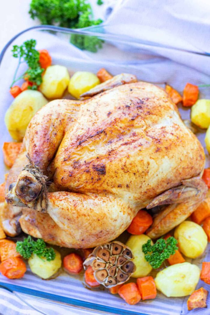 Roast chicken with crispy skin, thanks to a Whole Chicken Brine, surrounded by potatoes, carrots, parsley, and a halved head of garlic.