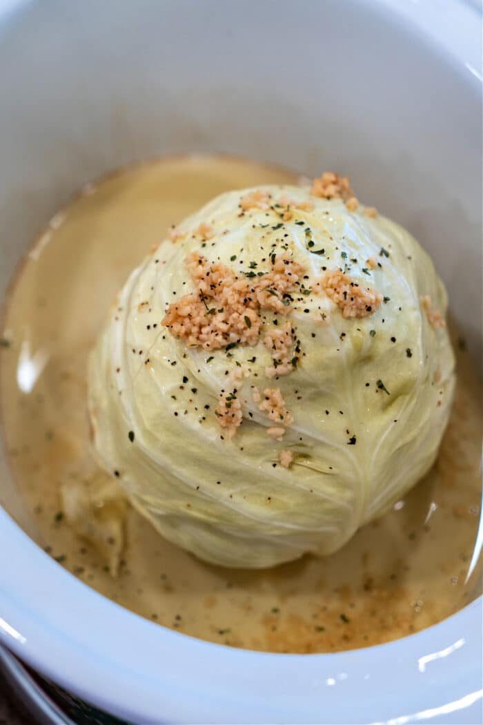A whole head of cabbage, slowly cooked to perfection in a white dish, is beautifully topped with minced garlic and herbs nestled in a savory broth.