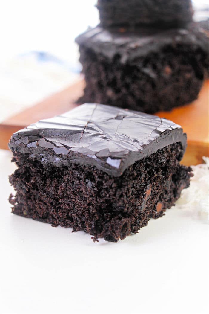 A close-up of a piece of chocolate cake with glossy, dark chocolate frosting, reminiscent of a rich zucchini brownie recipe, is placed on a white surface. Another piece of cake is visible in the blurred background on a wooden board.