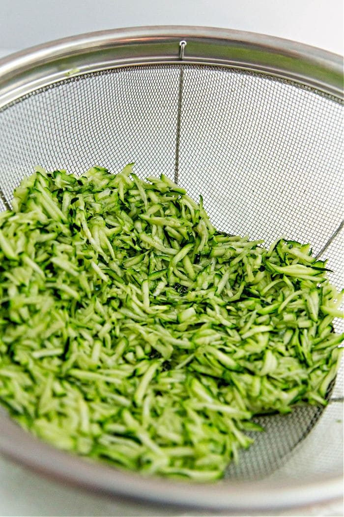 Shredded zucchini in a metal mesh strainer, angled thoughtfully. The green pieces are evenly distributed with visible moisture, ready to be transformed into crispy air fryer zucchini fritters. The blurred background enhances the focus on the vibrant veggies.