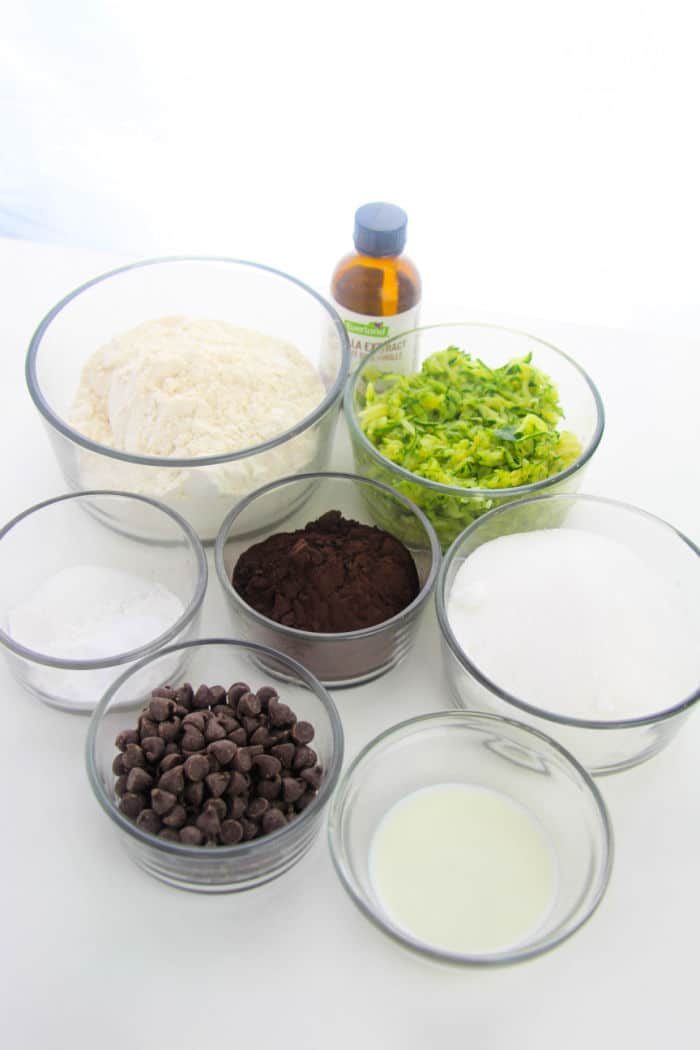 Seven glass bowls hold the ingredients for a zucchini brownie recipe, perfectly arranged on a surface. They include flour, chopped zucchini, sugar, chocolate chips, cocoa powder, a small bowl of liquid, and a bottle of vanilla extract.