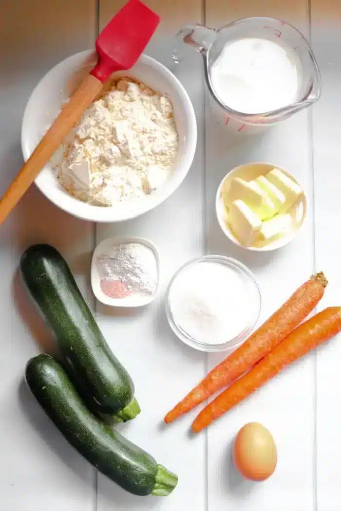A flat lay of ingredients for baking zucchini carrot muffins. Includes a bowl of flour, a red spatula, a measuring cup of milk, butter, two zucchinis, two carrots, a small bowl of salt and baking powder, a bowl of sugar, and one egg on a white table.