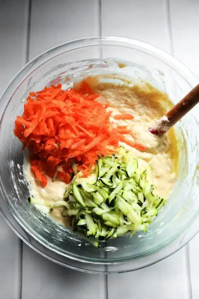 A glass bowl contains batter mixed with grated carrots and zucchini, solid for making good zucchini carrot muffins. A wooden spoon is partially submerged in the mixture, resting on a white countertop.