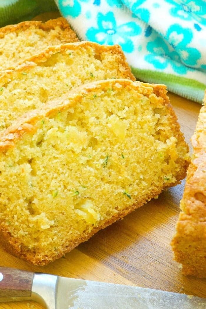 Slices of zucchini pineapple bread are displayed on a wooden surface. The bread boasts a golden crust and moist interior, with visible zucchini shreds and pineapple bits. A patterned green and white cloth graces the background, while a nearby knife awaits its next slice.
