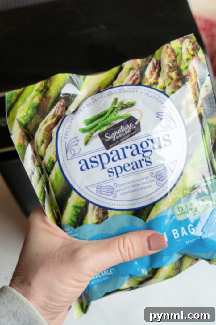 Crispy Air Fryer Frozen Asparagus 3 A hand holds a package of Signature Select frozen asparagus spears, perfect for the air fryer. The front label features images of asparagus and highlights product features such as being steamable and resealable.
