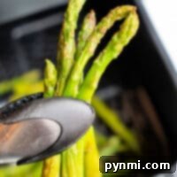 A close-up of a tong holding perfectly cooked, crispy asparagus spears over an air fryer basket captures the enticing appeal of these air-fried veggies.