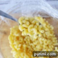A fork lifts cooked macaroni from a glass bowl covered with plastic wrap on a wooden surface, perfectly embodying the simplicity of how to cook pasta in the microwave.
