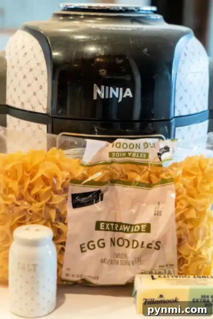 A sleek black Ninja Foodi air fryer looms in the background. Front and center, you'll find packages of egg noodles, a block of creamy Tillamook butter, and a chic white salt shaker adorned with gold dots—perfect ingredients for crafting delightful butter noodles.
