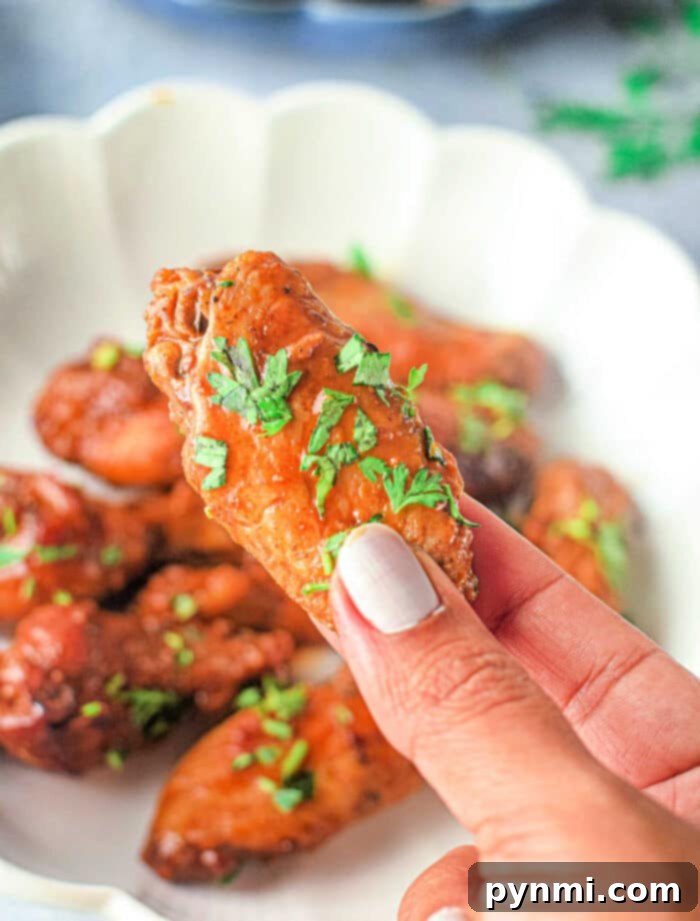 A hand holding a seasoned chicken wing garnished with fresh herbs over a white plate with additional Instant Pot Buffalo Wings in the background. The nails on the hand are painted with light-colored nail polish.