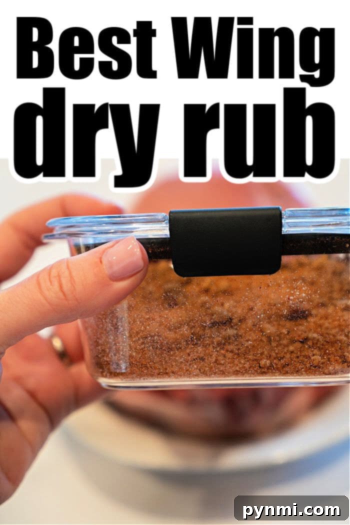 Master Griller's Dry Rub 5 A hand holding a glass container with a dark dry rub mixture perfectly crafted for chicken wings. The container, secured with a black latch on its lid, boasts the text: "Best Wing dry rub." In the blurred background, a plate subtly awaits the masterpiece.