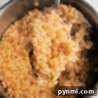 A spoon lifting cooked Israeli couscous over a bowl on a light background invites you to discover how to cook pearl couscous to perfection.