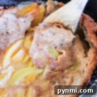 A close-up of a Crockpot cobbler with frozen fruit. A wooden spoon partially lifts the cobbler, revealing juicy sliced peaches and a golden brown, baked surface. The dessert looks moist and freshly cooked.
