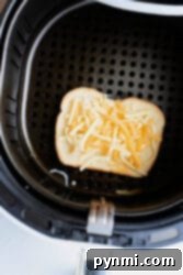 A slice of bread, generously topped with shredded cheese and a dollop of tuna mixture, sits inside an open air fryer basket, ready for its final crispy transformation into a tuna melt.