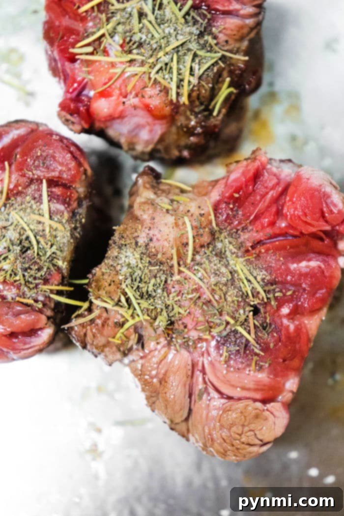 Stovetop Oxtail Perfection 4 Three raw pieces of oxtails, seasoned with herbs and spices on a metal surface. The herbs are visibly sprinkled on top, adding texture and color contrast to the red and pink hues of the meat.