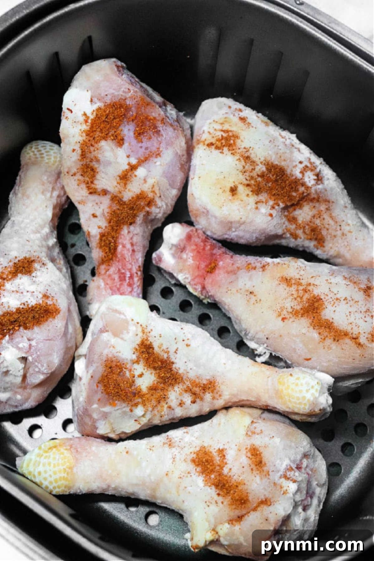Six raw, seasoned chicken drumsticks, perfectly positioned in an air fryer basket, awaiting cooking to become delicious Air Fryer Frozen Chicken Legs.