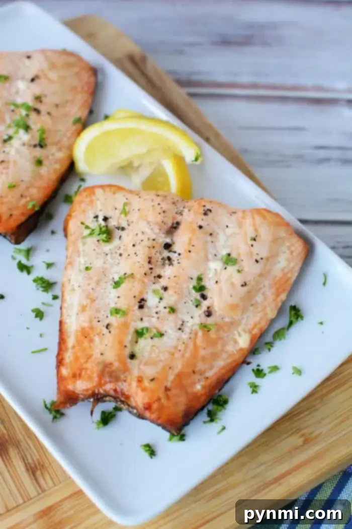 A delectable grilled salmon fillet rests on a rectangular white plate, garnished with chopped parsley and black pepper. Two lemon wedges accompany this delight. The plate, showcasing perfectly thawed frozen salmon grill markings, sits on a wooden cutting board with a striped cloth beneath.