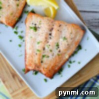 A grilled salmon fillet, previously frozen for freshness, glistens with chopped parsley on a white plate. Lemon wedges accompany it, all elegantly positioned on a wooden cutting board atop a plaid fabric.