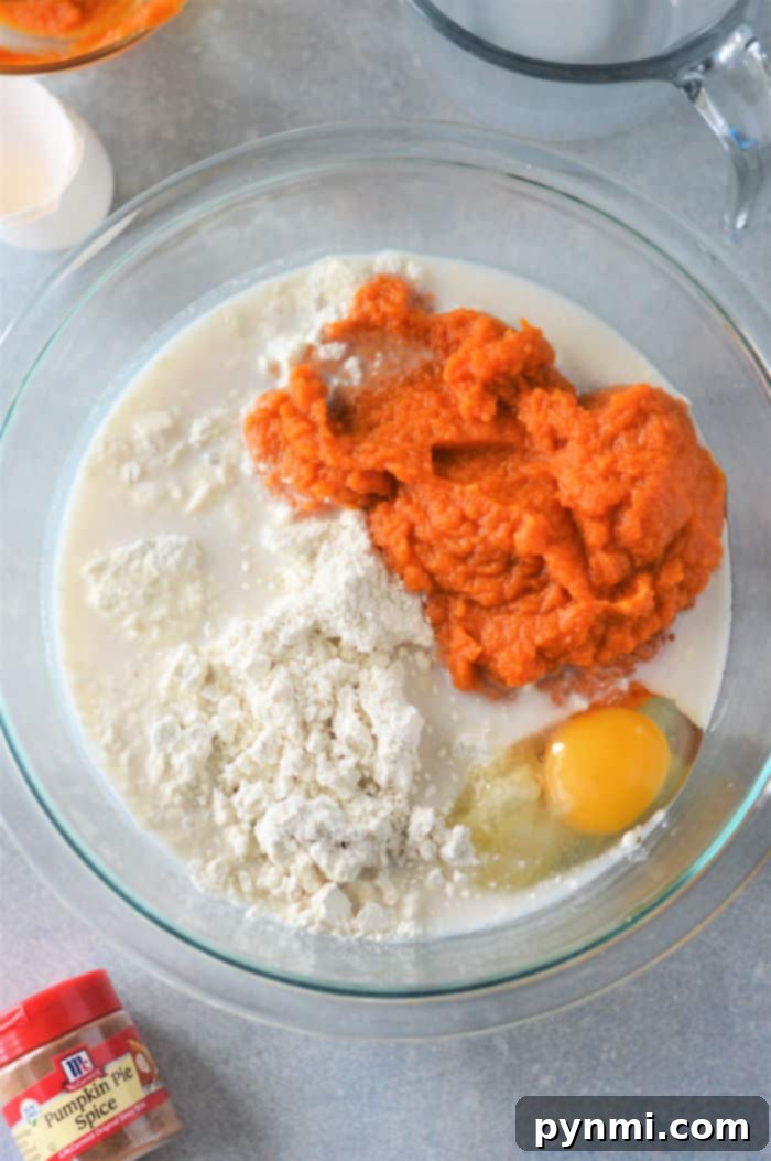 A mixing bowl filled with the base ingredients for pumpkin pancakes: Bisquick, pumpkin puree, a cracked egg, and milk. A container of pumpkin pie spice sits nearby, promising enhanced flavor.