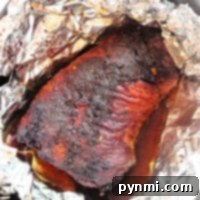 A cooked brisket is resting in a pan, wrapped partially with aluminum foil. The air fryer brisket appears well-seasoned and juicy, with a dark, caramelized exterior. There are flavorful juices collected at the bottom of the pan.