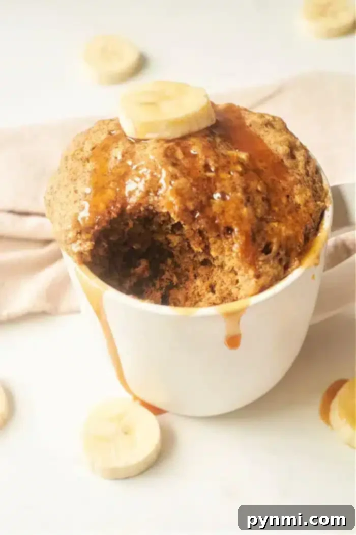 A Banana Bread in a Mug creation, topped with a banana slice and drizzled with syrup. The cake has a bite taken out of it, revealing its moist texture. Additional banana slices are artfully scattered around the mug on a light surface, enhancing its delicious appeal.