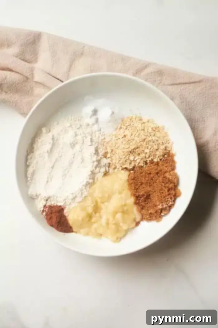 A white bowl holds the makings of banana bread in a mug: flour, oats, brown sugar, mashed banana, cinnamon, and a dash of baking soda. A beige cloth subtly peeks into view on the pristine white surface.