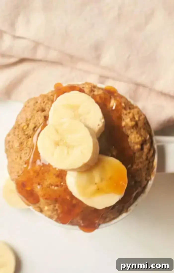 A banana bread mug cake topped with sliced bananas and drizzled with syrup is placed against a soft pink background. Some banana slices are also visible on the surface near the mug.